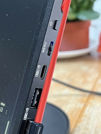 The image shows the side of a portable monitor featuring various ports, including a USB-C port, a USB-A port, buttons for power and a rocker switch for menu functions. The device has a sleek design with a an orange red flip cover just visible behind it.