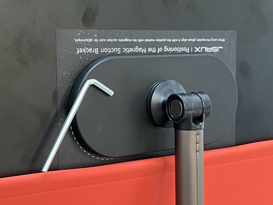 An adjustable mounting bracket with a black and silver finish, accompanied by an L-shaped tool magnetically stuck to it. The bracket appears to be affixed to the back of a portable monitor with an orange red flip cover hanging below.