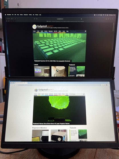 A dual-monitor setup displaying the Gadgetoid website. The top monitor features a green-themed article about a keyboard, while the bottom monitor features an article on a 4K laser projector. The top website is Firefox dark mode, and the bottom is Chromium in light mode. The light is… noticeably dim. A wooden table is visible in the background.