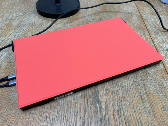 A portable dual 15.4” monitor closed on a tabletop. It’s protected by a bright reddish orange flip cover. USB type C cables snake out of the left and a tabletop stand is just visible in the background.