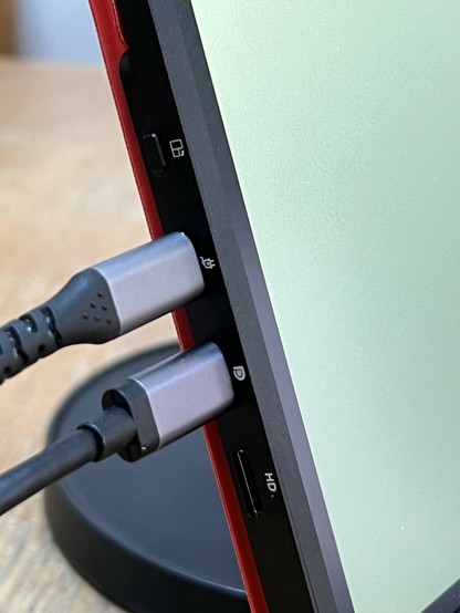 Close-up of the side of a device with two cables connected to ports labeled with icons. The ports include a USB-C, HDMI, and power. Theres a button with a weird icon that looks like it’s for rotation.