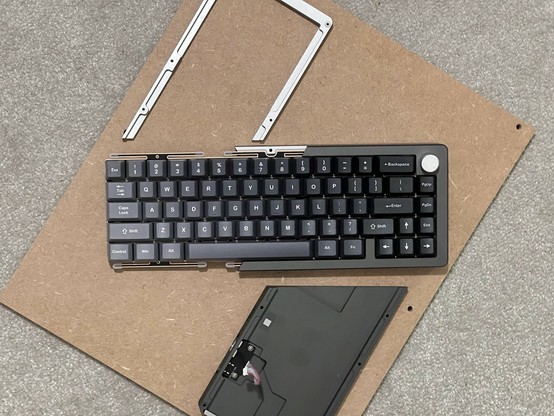 A disassembled mechanical keyboard is displayed on a small sheet of MDF. Half of the keyboard outer shell has been removed and set aside almost as if it has been cut diagonally. It’s just… built that way.