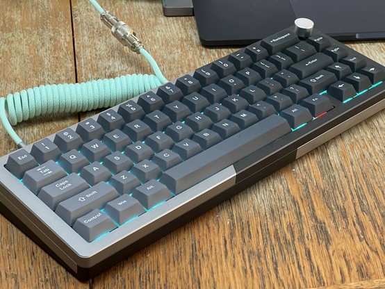 A mechanical keyboard with a sleek design, featuring dark gray keys illuminated by a teal glow. The keyboard is positioned on a wooden surface, with a coiled mint green cable visible. A laptop is partially visible in the background.