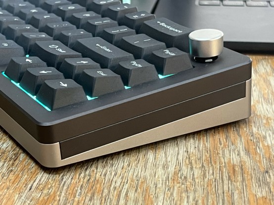 An angled view of a mechanical keyboard with backlit keys, featuring a metal volume knob on the side. The keyboard is about an inch thick at its thickest and the edge on view shows three separate metal parts.