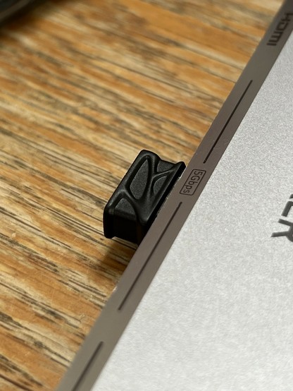 A close-up image of a small black USB dongle plugged into the side of a metallic USB hub, resting on a wooden surface. The clip appears to have a textured, alien design.