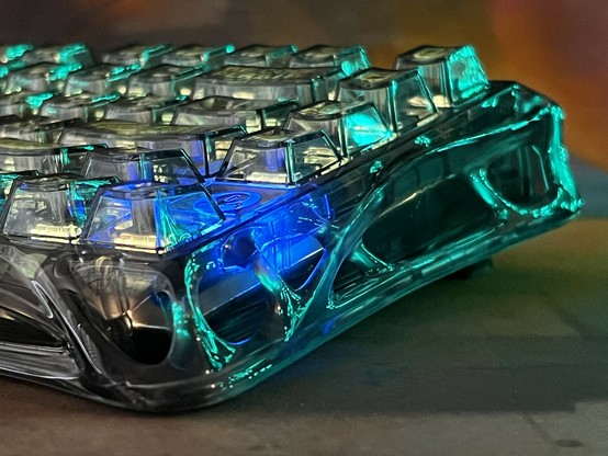 A close-up view of a translucent mechanical keyboard with colorful backlighting, showcasing the intricate design of the transparent keys and a glowing (with reflected light), sculpted, alien-looking base.