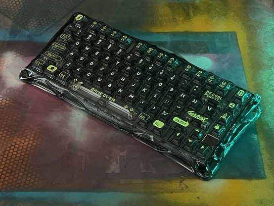 A black gaming keyboard with white and yellowish green legends on transparent keys displayed on a colorful, patterned surface. The keyboard features a 