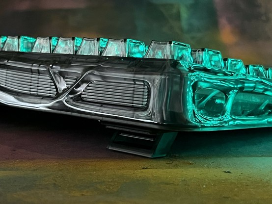 A close-up view of a transparent, alien looking mechanical keyboard with glowing teal lights reflected in the top and sides. It’s raised off a textured, colourful painted surface with a fold out foot.