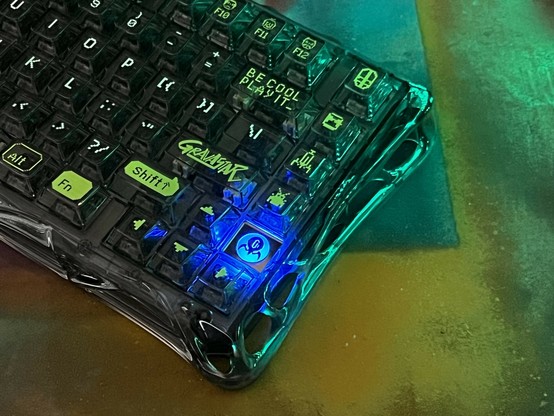 A close-up of a translucent black keyboard with key legends picked out in white and yellow green. The keyboard features distinct branding and pixel art icons on some keys. The background has a colorful, abstract texture. (It’s my spray paint overspray cardboard)