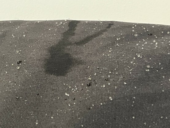 A textured black fabric surface with beads of water droplets and a distinctly dark wet mark where the brunt of the water hit and ran off the edge.
