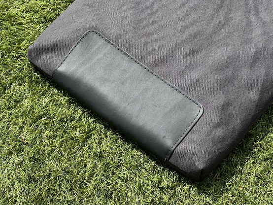 A close-up of a black fabric sleeve case (though it looks grey) with a black leather patch folded over the bottom, resting on green artificial grass.