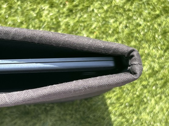 A blue laptop inserted into a grey fabric casing, placed on green grass or turf. The case is open showing the laptop inside and the thickness of the padding.