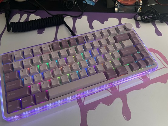 A mechanical keyboard with a compact layout, featuring purple and off white colored keys and multicolored RGB backlighting. The keyboard is mounted on a translucent base, and there is a coiled cable visible in the background. The setup is placed on a colorful, drippy desk mat that matches.