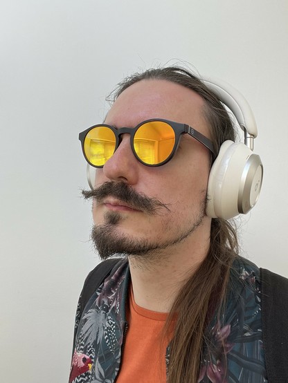 A white moustachioed male wearing some orange lens sunglasses and a pair of white headphones. Looks somewhat unkempt.