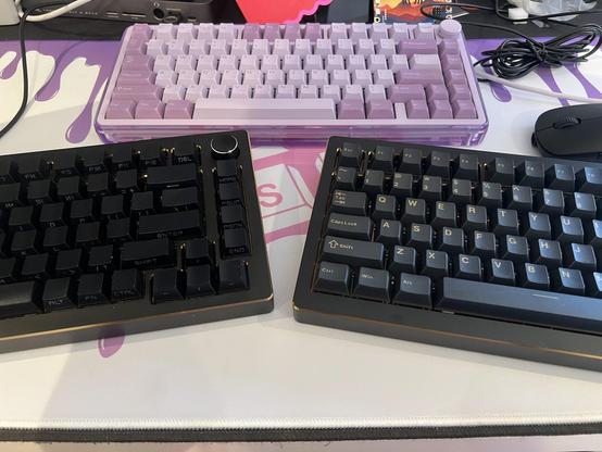 Three keyboards on a colorful desk mat. Two are black with a sleek design and gold accents, while the other is light purple. Each has a compact 75% layout.