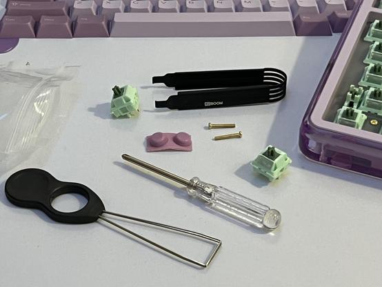 Keyboard parts and tools, including green switches, a screwdriver, a pair of switch pullers, and screws, arranged on a light surface with keycaps lined up in the background.