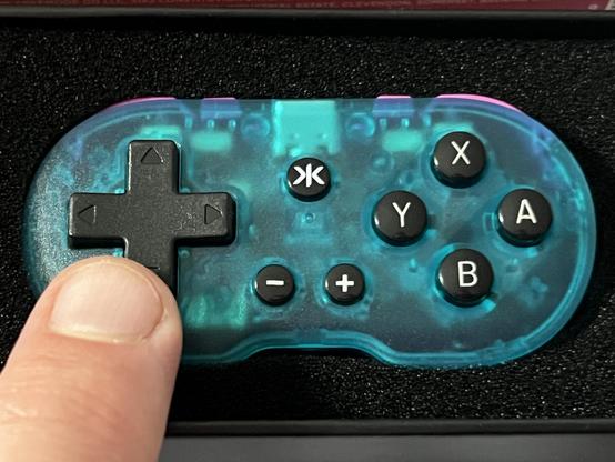 A translucent teal and pink game controller with a directional pad, four action buttons labeled A, B, X, Y, and a start, select, and +/- buttons. A finger is pressing the directional pad revealing the controller to be adorably tiny.