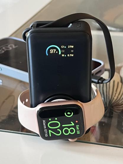 A smartphone/watch charger with a little LCD display showing charge level and output power. An Apple Watch is wrapped around it and charging. A phone sits in the background, connected via a captive USB cable.