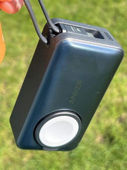 A black and gray portable charging device with a circular Apple Watch charging pad, held in the air by its captive USB cable.