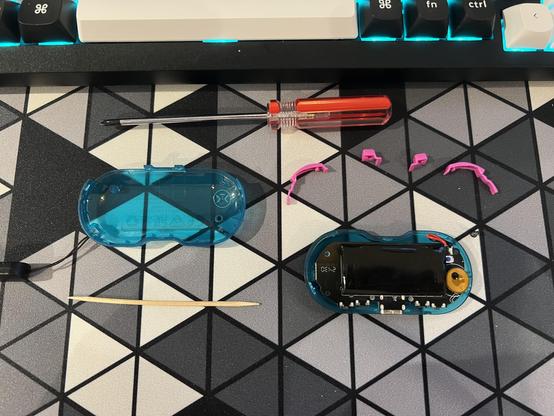 A disassembled game controller device featuring a transparent blue casing and an internal circuit board mostly obscured by battery. Nearby are a small screwdriver, a cocktail stick, and pink plastic shoulder buttons.