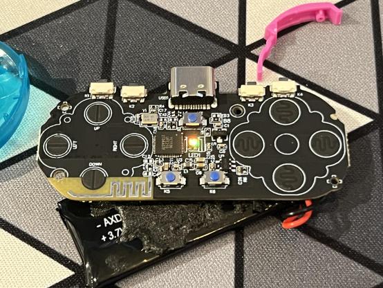 An tiny gamepad circuit board with 
a USB-C port, and a battery attached. Three tactile switches with blue buttons are arranged in the middle and a small light is illuminated between them. Four right angle tactile switches are arrayed across the top edge for shoulder buttons. Eight PCB switch contacts for rubber dome switches are arranged into a D-Pad and face button configuration on each side.