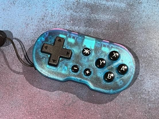 A translucent blue gaming controller with a black directional pad and buttons, set against a textured blue and pink painted background.