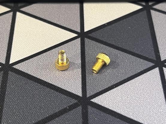 Two small brass screws are positioned on a patterned grey desk mat. They are short, with large, grippy heads.