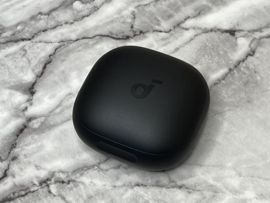 A small matte black, rounded pebble-like Soundcore earbuds case on a marble textured surface.