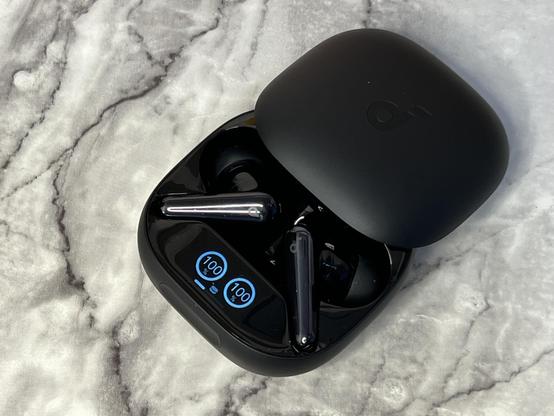 A pair of black wireless earbuds in their charging case, resting on a marble textured surface. The case displays battery status indicators showing 100% charge for both earbuds.