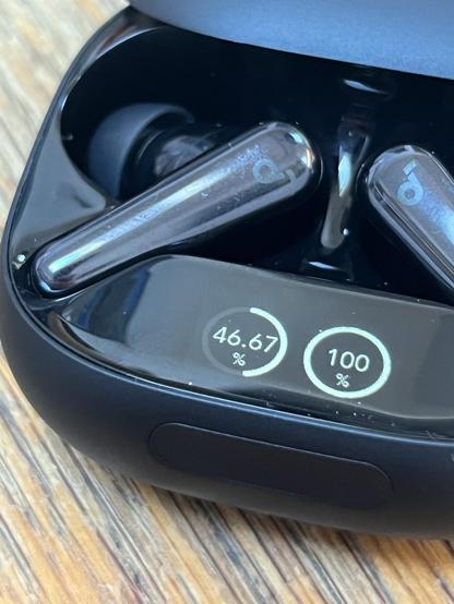 A charging case for wireless earbuds, displaying battery percentages: 46.67% for one earbud and 100% for the other. The case has a sleek black design with a glossy internal finish. The earbuds are glossy and metallic.