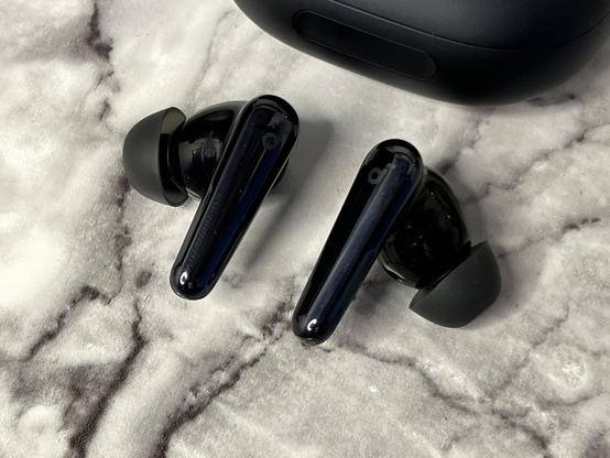 Two  glossy metallic black wireless earbuds with a charging case in the background, placed on a marble textured surface.