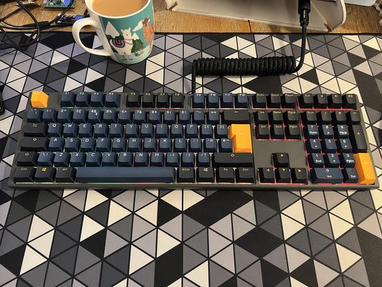 A mechanical keyboard featuring dark blue, black and orange keys. It is placed on a geometric-patterned desk mat, alongside a mug of tea. The keyboard has an (aftermarket) braided cable.