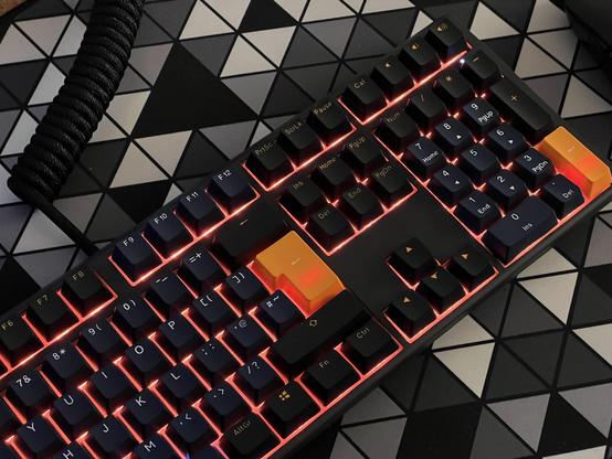 A mechanical keyboard with grey and black keycaps. The Enter and NumPad Enter keys are accented in orange. The orange backlighting shines around the keycaps and creates little hotspots behind the orange keys.