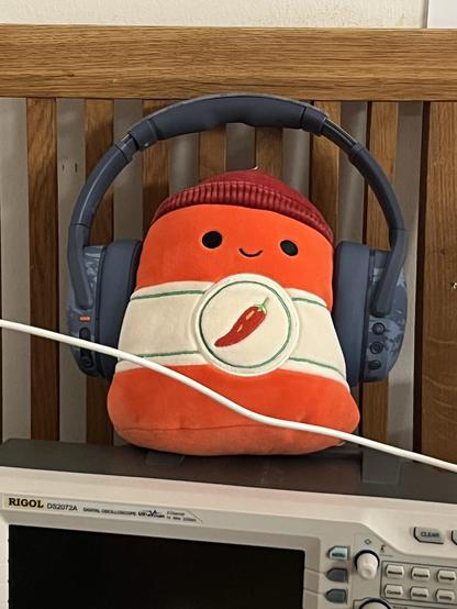 A chilli sauce squish mallow plushie with a pair of blue on ear headphones over it. It’s sat on top of an oscilloscope on a dining chair. I am chaos.