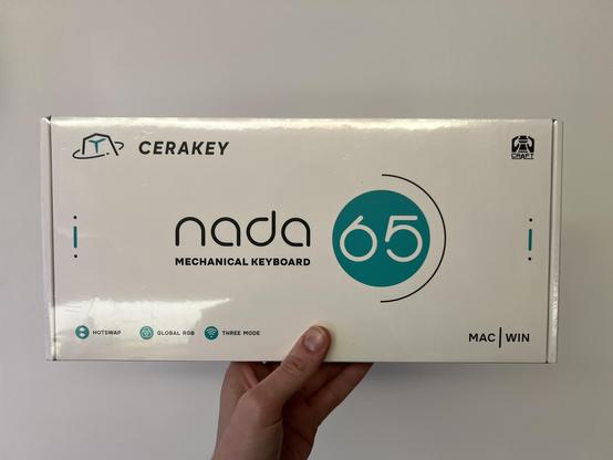 An individual holding a box for the Cerakey Nada 65 mechanical keyboard. The box features a white design with a teal circle and text indicating 