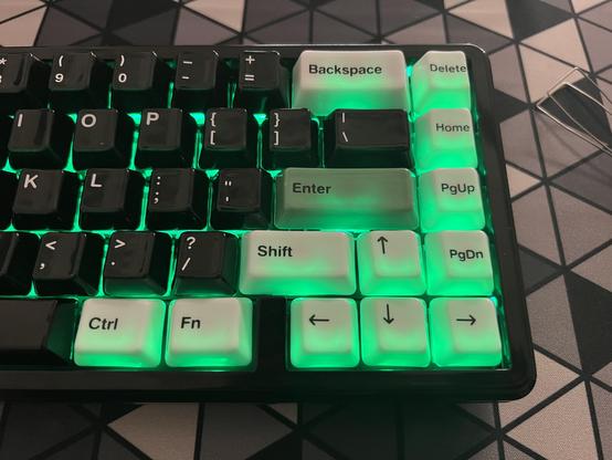 Closeup of white and black keycaps lit up by a green underglow. The white caps are glowing green, but the very top edges of the black caps seem to be too.