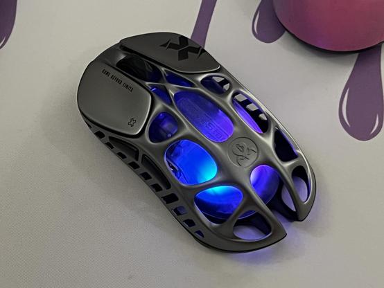 A silver mouse on a white desk mat. The mouse has a skeletal, lightweight outer shell revealing a glowing RGB LED lit inner shell.