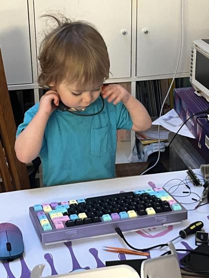 A purple mechanical keyboard with black and pastel keycaps sat on a cluttered desk. A small child sits at the desk, putting on some bone conducting earphones ready for a hard days hyperfocus.