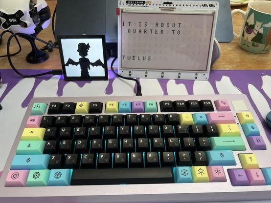 A icky role mechanical keyboard with black and pastel keycaps sat in front of an e-ink word clock and a square display showing a black and white silhouette of an anime bat girl. (Bad Apple, the hello world of displays and video decoding)