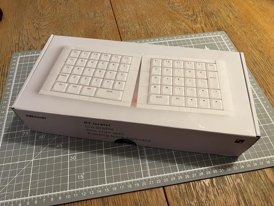 A KBDCraft Israfel “Lego style” split mechanical keyboard kit box, sitting on a cutting mat on a worn dining table.