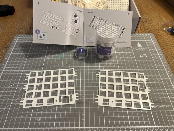 Two white ortholinear keyboard plates sat on a cutting mat. Behind is a box of switches and an assembly instruction manual.