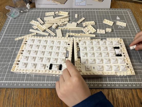 The PCBs inserted into the two keyboard halves which have been joined by a hinge. A small hand is adding bricks.