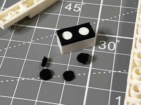 A sticky foam pad stuck over the studs of a stack of two 1x2 LEGO plates. Some torn and mangled bits - removed to accommodate the studs- sit alongside, suggesting some trouble removing them.