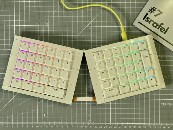 An overview of the finished keyboard, a white, Ortholinear, ergo split sat on a grey cutting mat and illuminated with rainbow backlighting that peeks between the keys.