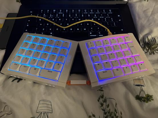 A split keyboard now tweaked with macOS keycaps. Sat on top of a MacBook on a bed.