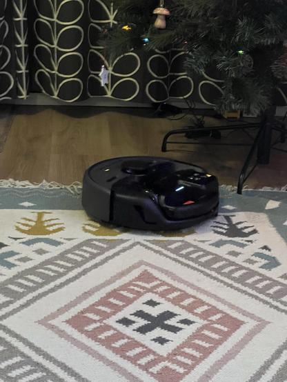 A robot vacuum cleaner approaching the edge of a tasseled rug at the base of an artificial Christmas tree.