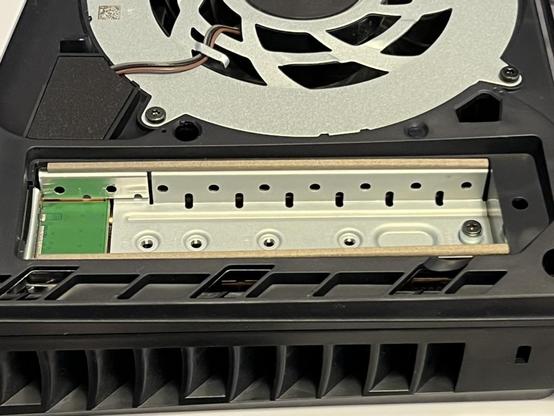 The SSD recess in the Playstation 5 slim. It's a metal cage sort of thing with a green PCB at the left where the NVMe slot sits. There appears to be some accommodation for airflow with small holes in the sides, but not much. Four empty screw holes run across the bottom with a fifth occupied by a riser and screw.