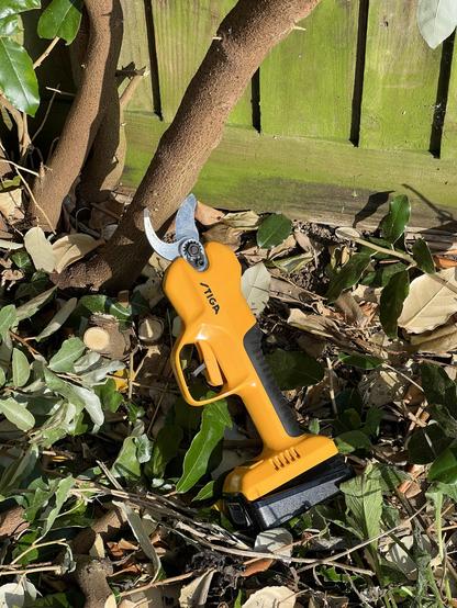 A pair of yellow electronic pruning shears lying on the ground with their jaws posed around a branch too big to even fit within them.