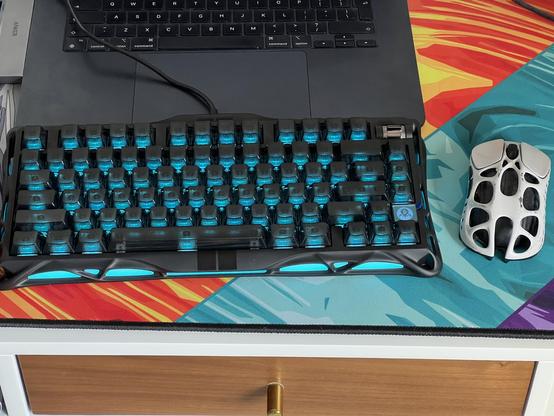 A black organic alien styled mechanical keyboard sat next to a mouse in a similar style. They are both perched in front of a very not gamer MacBook Air.