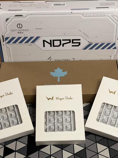 A boxed ND75 mechanical keyboard with a box of keycaps in front of it, adorned with a maple leaf logo. Three packs of white Wuque Studio switches lean against the keycap box. It’s all the bits for a mechanical keyboard build.